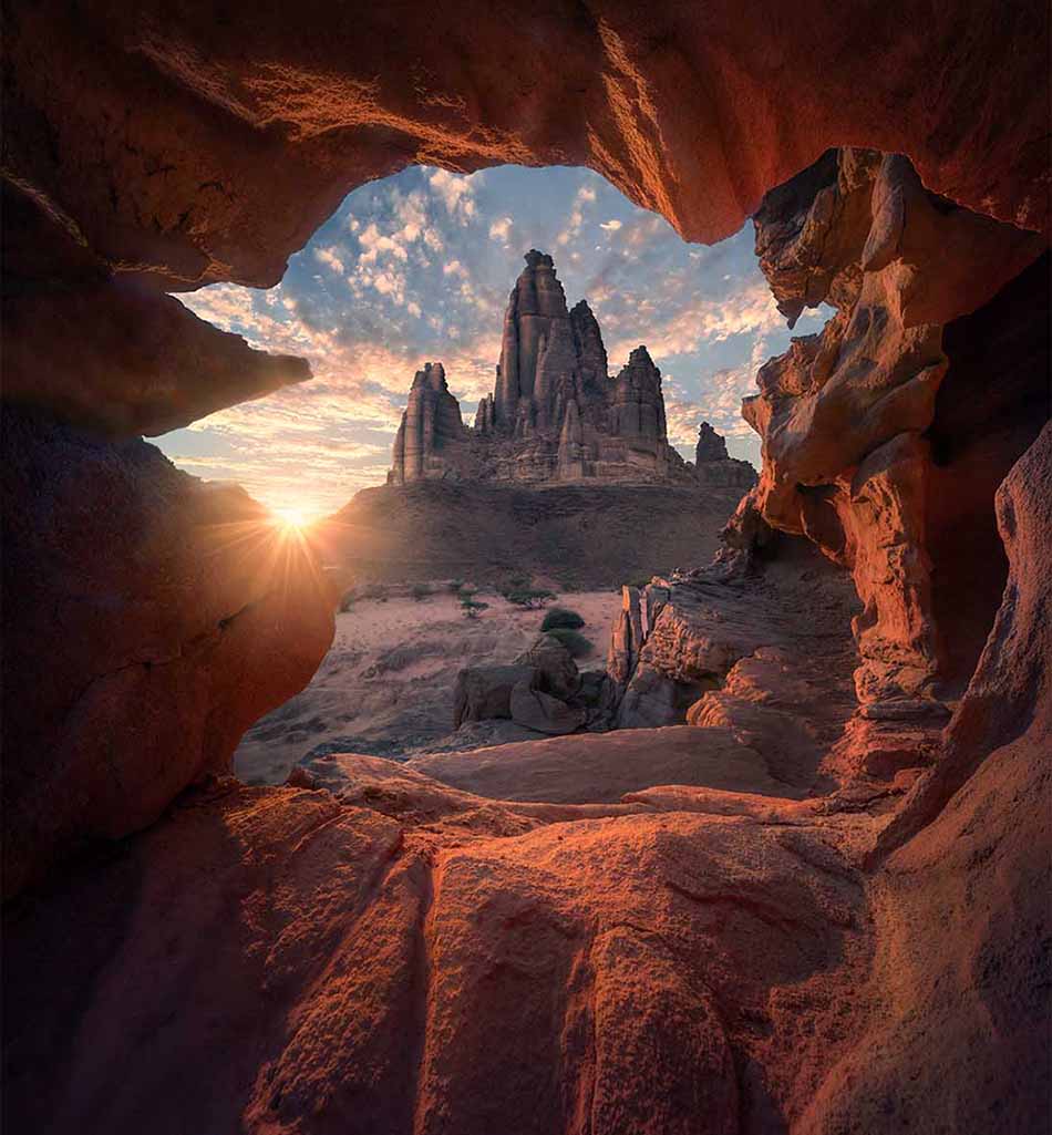 A dramatic landscape photograph of a desert rock formation seen through a natural cave opening at sunset, with warm orange and red sandstone framing the scene and a bright sun flare on the left.