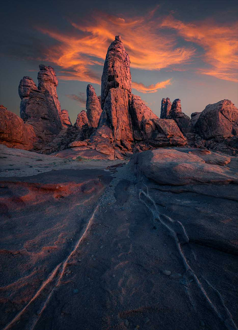 Red light on sandstone spires beneath orange clouds, with smooth rock in the foreground in Saudi Arabia.