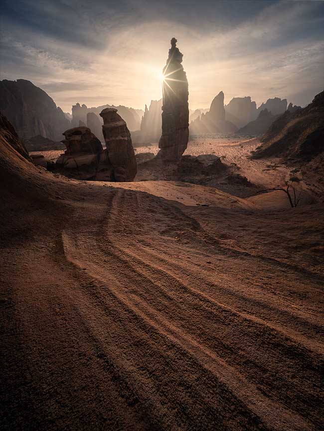 A dramatic desert landscape with a tall rock spire silhouetted against a low sun, casting strong backlighting across a sandy valley and distant rock formations.