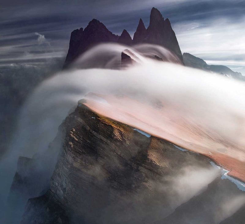 The mountain peaks of Seceda emerging from a cloud blanket with the ridgeline emerging from the mist. The waterfall of clouds goes over the ridge but is beneath the peaks.