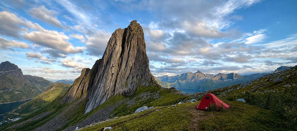A red tent sits on a grassy hill overlooking Segla mountain in Norway during the midnight sun. Soft light illuminates the peaks of the surrounding fjords and mountains.