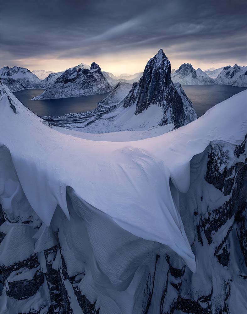A sharpy snow banked ridgeline above Segla, taken by drone in winter catches the sunrise light. Behind the sharp fin of segla mountain dominates the scene with the golden sunrise reflecting in the glassy fjords below.