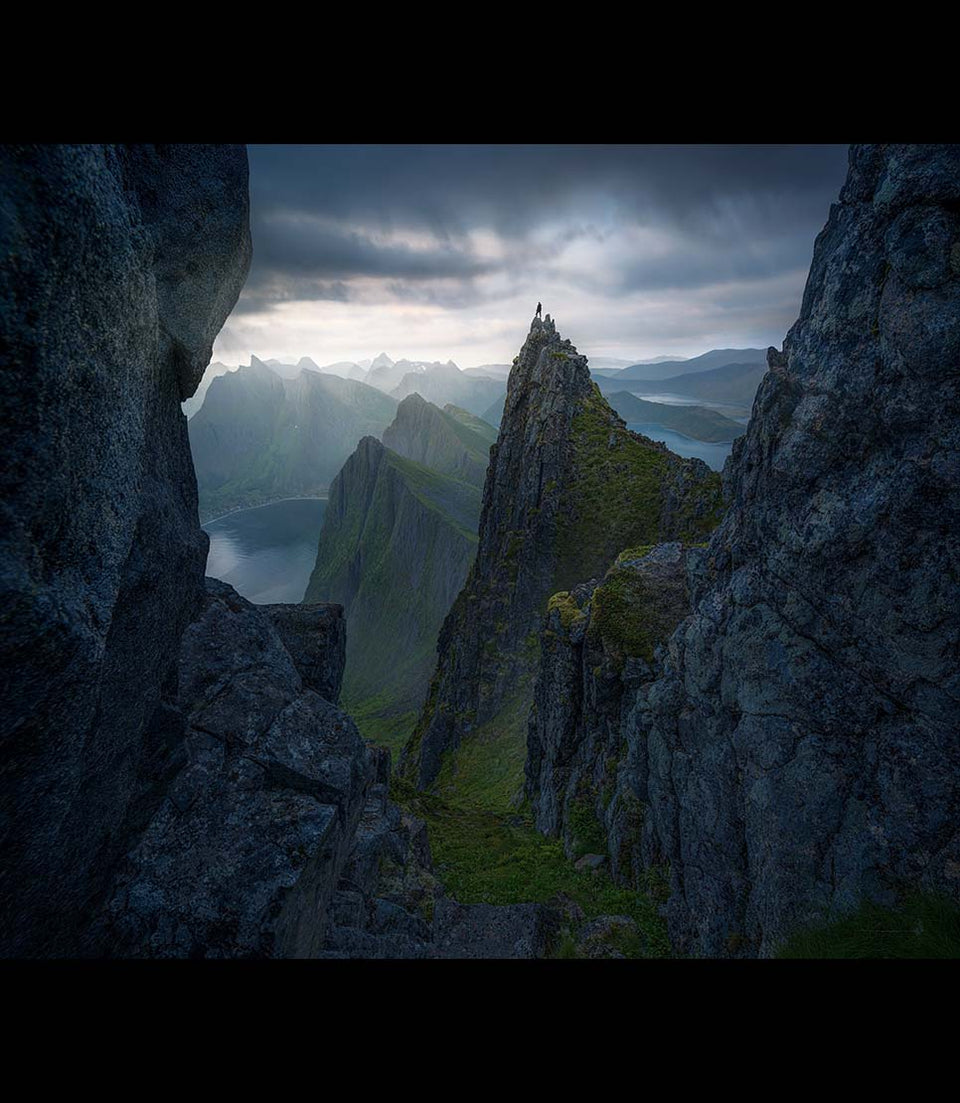 A person stands on the edge of sharp peak in Norway overlooking the fjords below, the sky is a dramatic stormy grey and the peak is framed by two rocky formations.