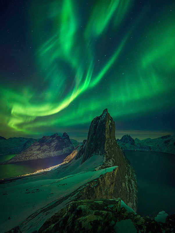 segla mountain in Norway in winter. The northern lights are bright green and luminescent above the peak. Either side the fjords frame the mountain.