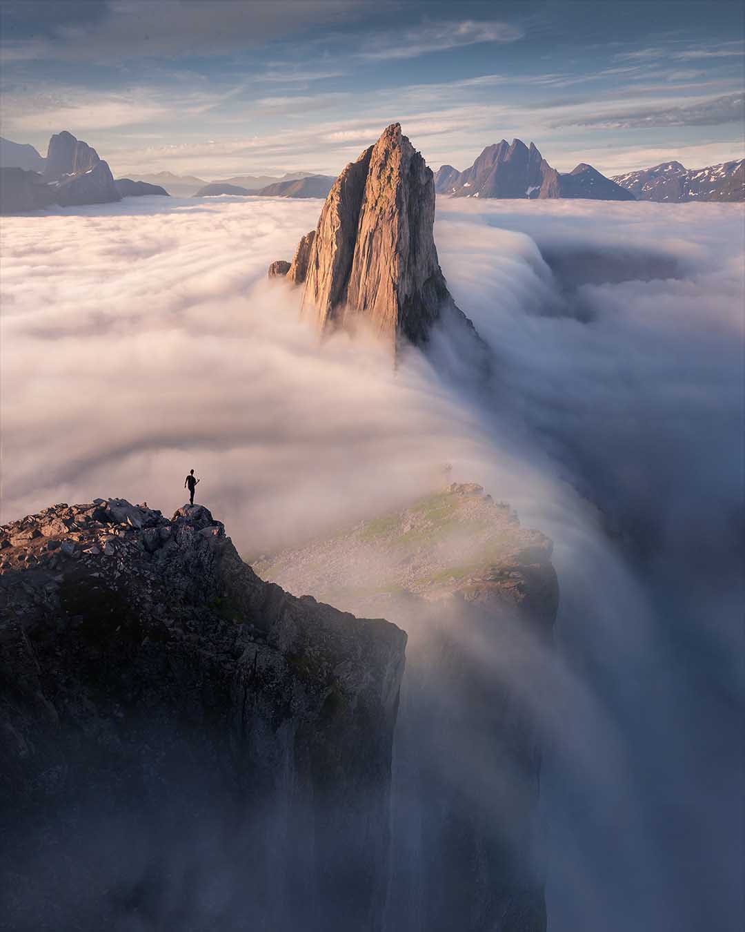 A dramatic mountain landscape with a tall rocky peak rising above a sea of clouds, with a small human figure standing on a cliff edge in the foreground. The scene is lit by soft golden light on the rock and cool blue light on the surrounding mountains and mist.