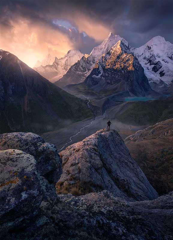 man stand on the edge of a rock high in the peruvian andes looking at the mountains at sunset. in the valley below is a blue glacier lake and a winding river and waterfall.