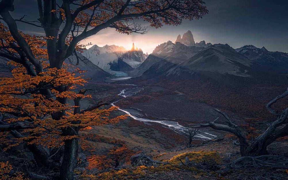 Cerro Torre in Patagonia seen from a framing of autumn coloured trees. sunrays spread from the mountains over the valley. a river creates a leading line to the background.
