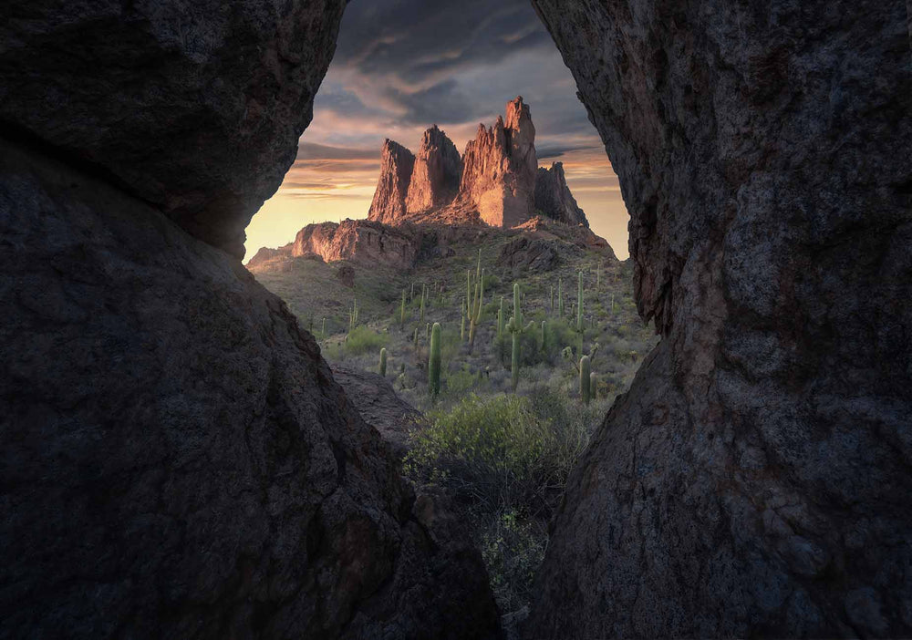 superstition mountains at sunset through a rock cave which frames the mountains. The golden and orange light glows on the mountain. Beneath the rocky formations a field of cacti stand tall.