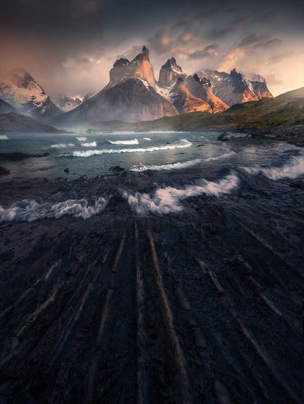 The rugged mountain peaks of Torres del Paine in Patagonia rising above a dark rocky shoreline with foamy surf in the foreground, under a cloudy sky lit by warm light near the mountain peaks.