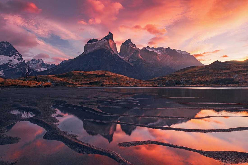 Torres del Paine in Chile, Patagonia at sunrise. The sky and mountains are bright pink with colour and are reflected in the sandy shoreline of the lake below.