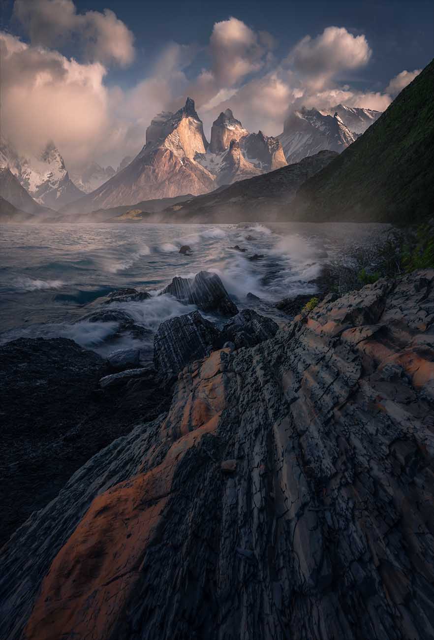 Torres del Paine jagged mountain peaks rising above a rocky shoreline of Lake Pehoe, with waves crashing in the foreground and clouds filling the sky, orange lines in the rocks lead into the shoreline.