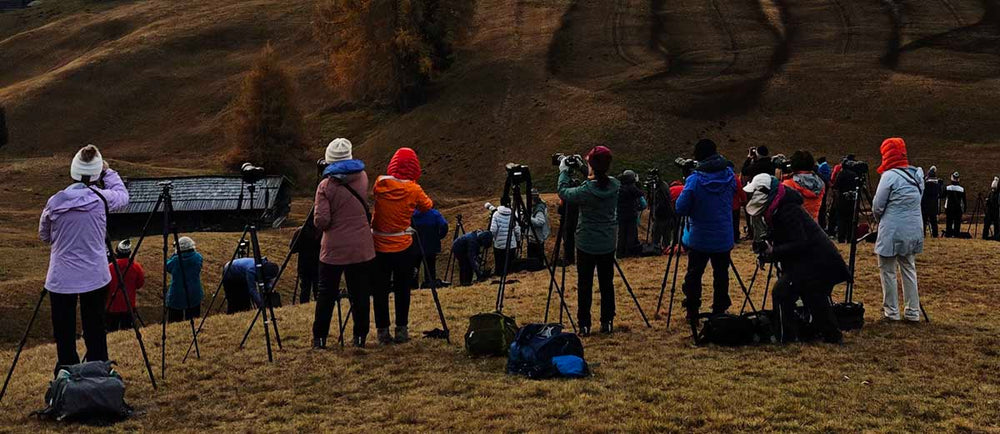 A large group of photographers stand on a grassy hilltop in soft morning light. Every person has a tripod. Some camera bags lay around on the grass.