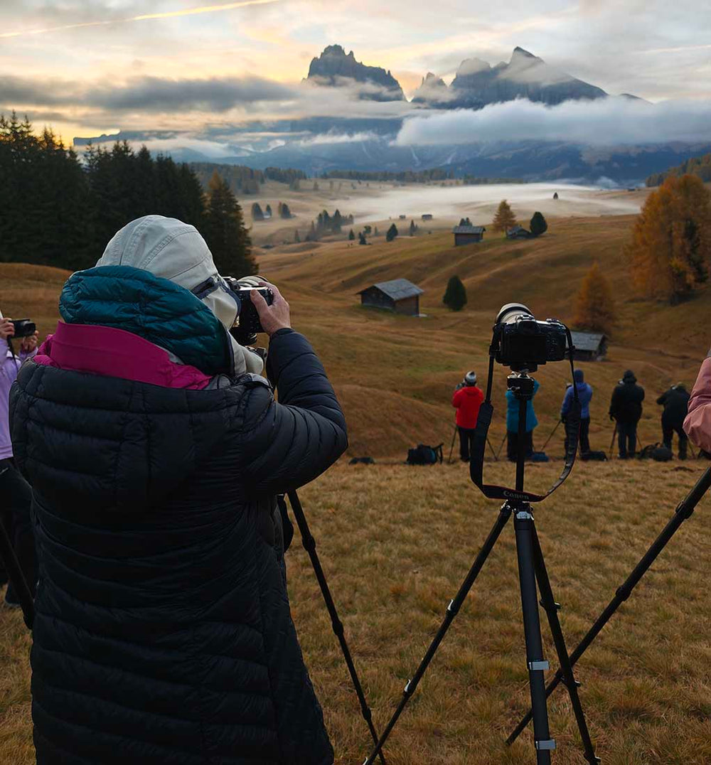 A person stands with their tripod next to two other tripods on a grassy bank. In the distance are the mountains of alpe di siusi shrouded with mist. In the midground the trees and huts stand out amongst the layers of mist.
