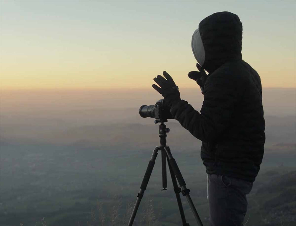 a man stand with a tripod and camera at sunset overlooking a misty valley.