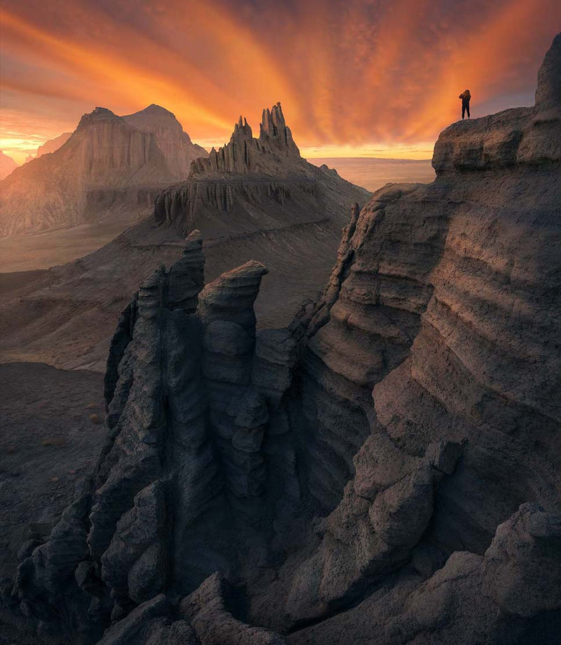 A castle shaped rock formation in the kazakhstan desert, a lone figure stands on the edge of the ridge looking into the firey orange clouded sky, there are lines of rock running along the edge of the cliff creating depth.
