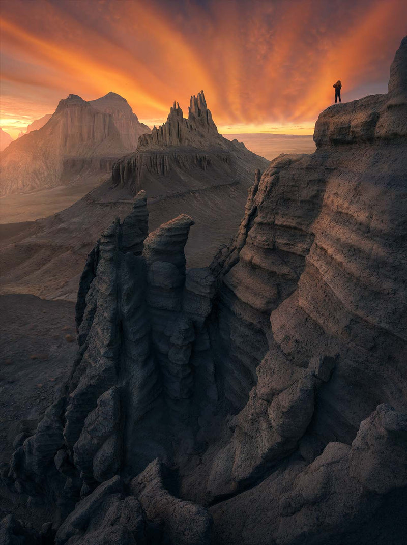 A lone figure above layered cliffs and sharp rock towers beneath an orange sky in Mangystau.