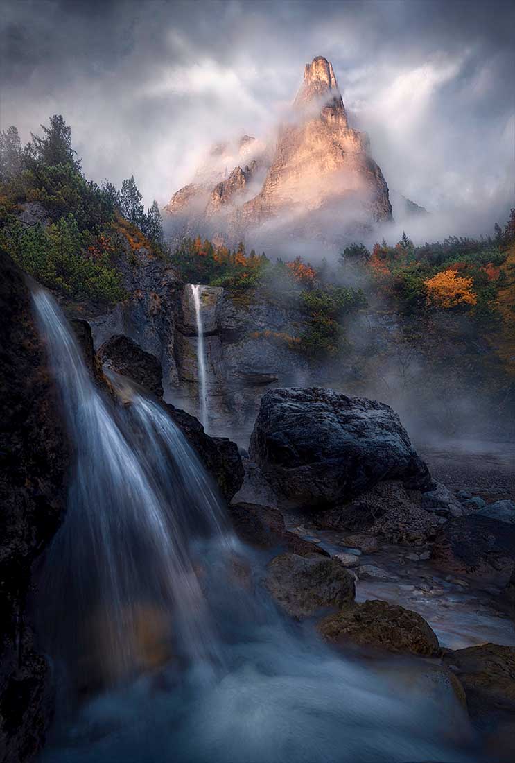 fall coloured trees stand above a high waterfall. Misty mountains are high in the background.