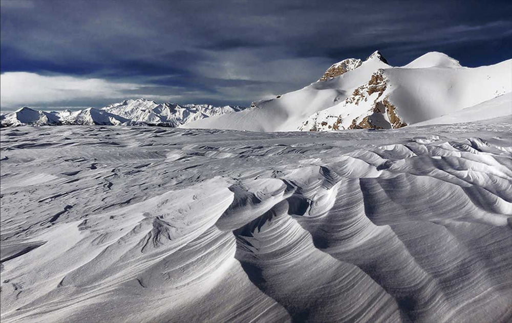 Snow patterns caused by gusts of wind create leading lines in the ridges of a snow field. In the background a large snow covered mountain dominates the landscape.
