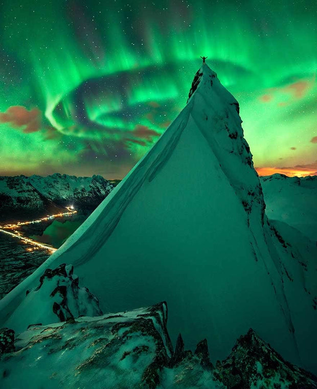 Green Colored snowcapped mountain during northern lights with person standing on top
