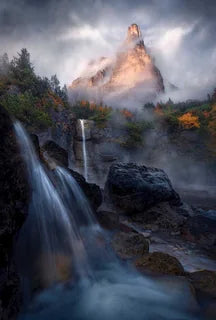 A tall castle shaped mountain peak in the dolomites surrounded by clouds and mist catching the last golden light, an autumn coloured forest beneath the peaks with two tall waterfalls in the foreground.