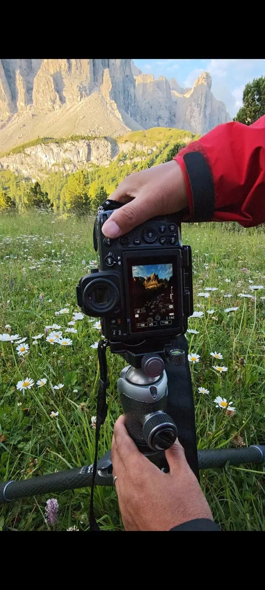 Hands adjusting a camera on a tripod among white wildflowers beneath sunlit cliffs in the Dolomites