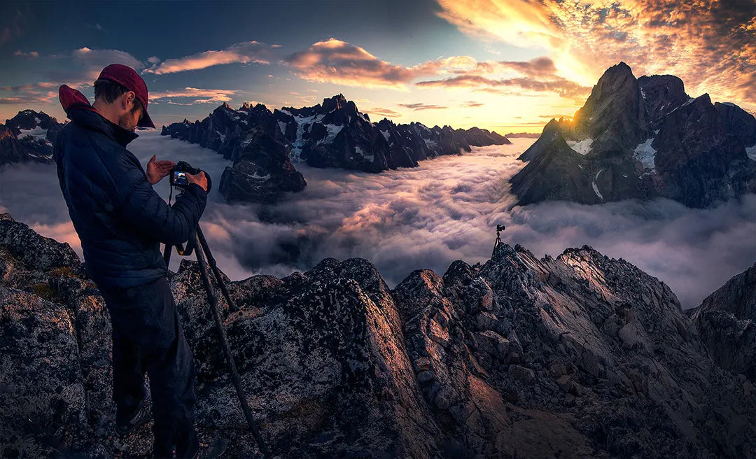 A photographer adjusting a camera on a rocky ridge above clouds and mountain peaks at sunset