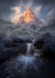 A dramatic mountain landscape with a tall rocky peak rising through mist, a small waterfall flowing over dark rocks in the foreground, and clouds around the summit, a dense forest with autumn colours is shrouded in mist beneath the mountain peaks.