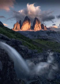 A dramatic mountain landscape of Drei Zinnen, a large waterfall in the foreground from the left flows right and towards the peaks, green slopes, and clouds lit by warm light near the peaks.