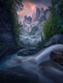 A majestic mountain landscape in the Dolomites in summer with a waterfall cascading towards the mountain peaks framed by tall green trees on each side. The clouds glows pink and mist streams through the valley and around the peaks.