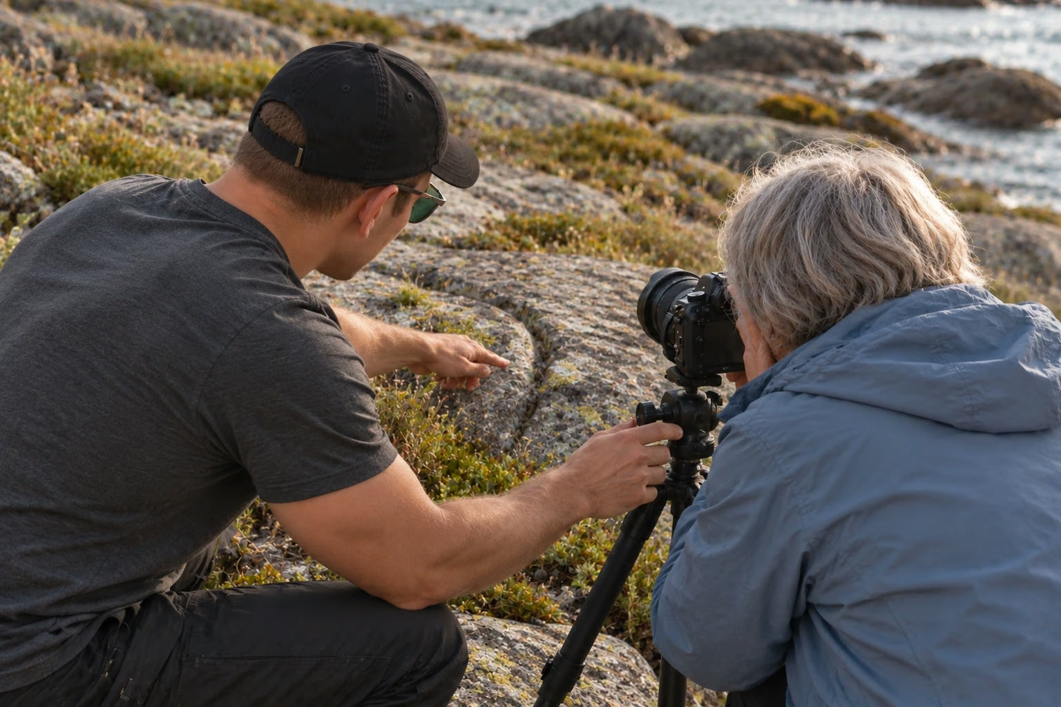 An instructor helping a photographer use a tripod-mounted camera beside coastal boulders in Senja