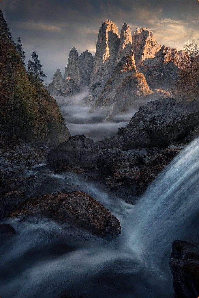 A waterfall flowing past dark rocks below sunlit peaks and low mist in the Dolomites.