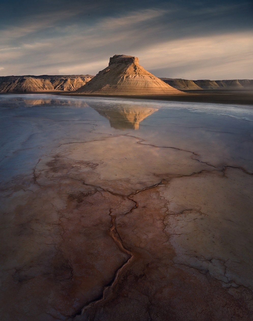 Warm light on a flat-topped hill reflected in shallow water above cracked ground in Mangystau.