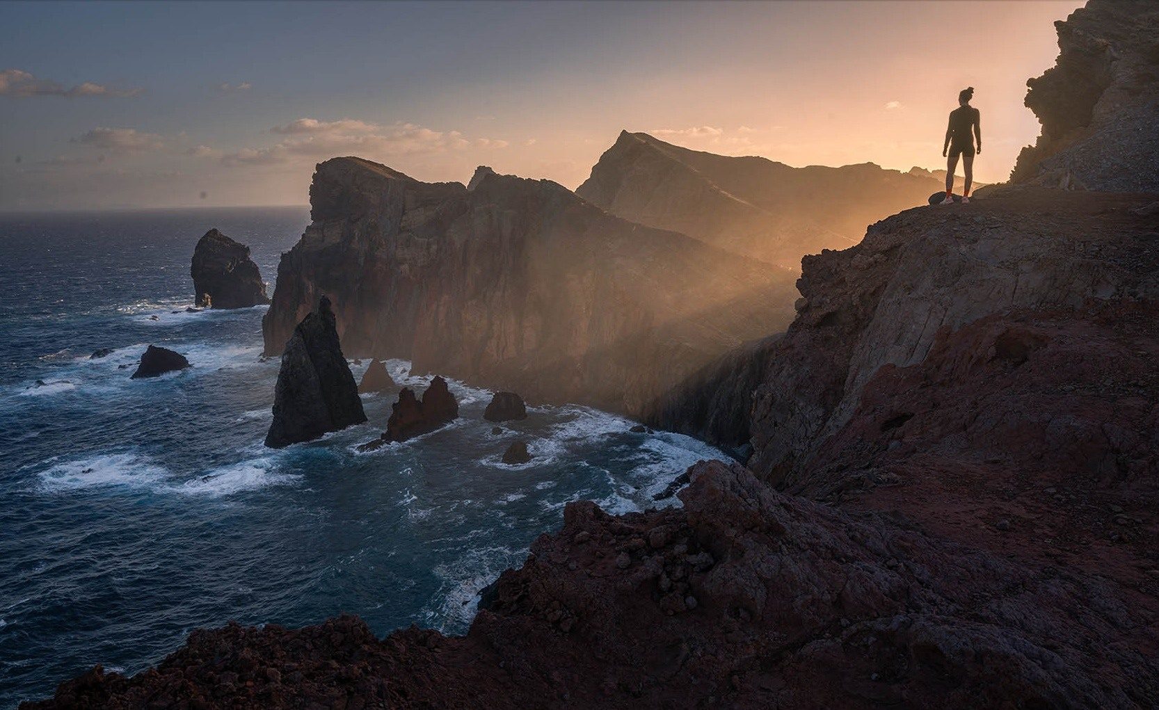 A hiker on a rocky headland above Madeira's sea cliffs in low golden light.
