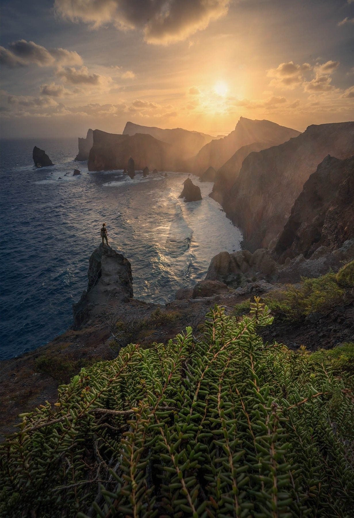 A small figure on a rocky coastal pinnacle above the Atlantic, with sunlit cliffs beyond on Madeira.