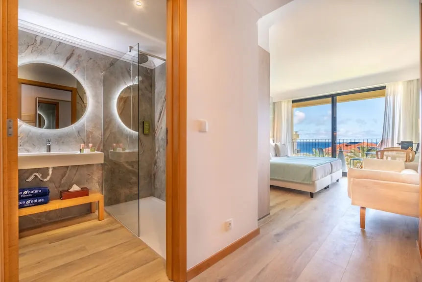 A bright hotel room with an adjoining bathroom and a balcony overlooking the sea