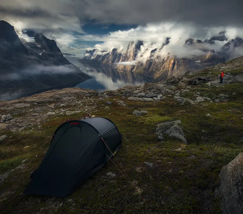 A tent on a rocky mountainside overlooking a fjord beneath dramatic clouds
