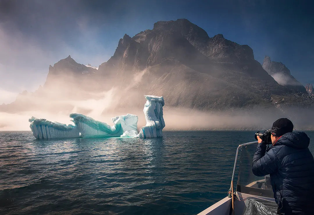 A photographer on a boat capturing a blue iceberg beneath misty mountain peaks