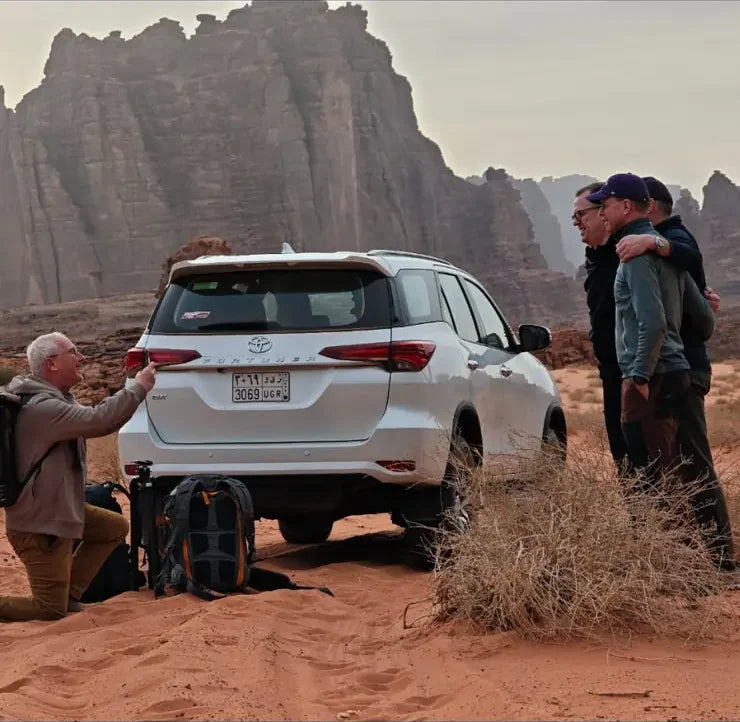 Friends posing for a photograph beside a white four-wheel drive beneath desert rock formations