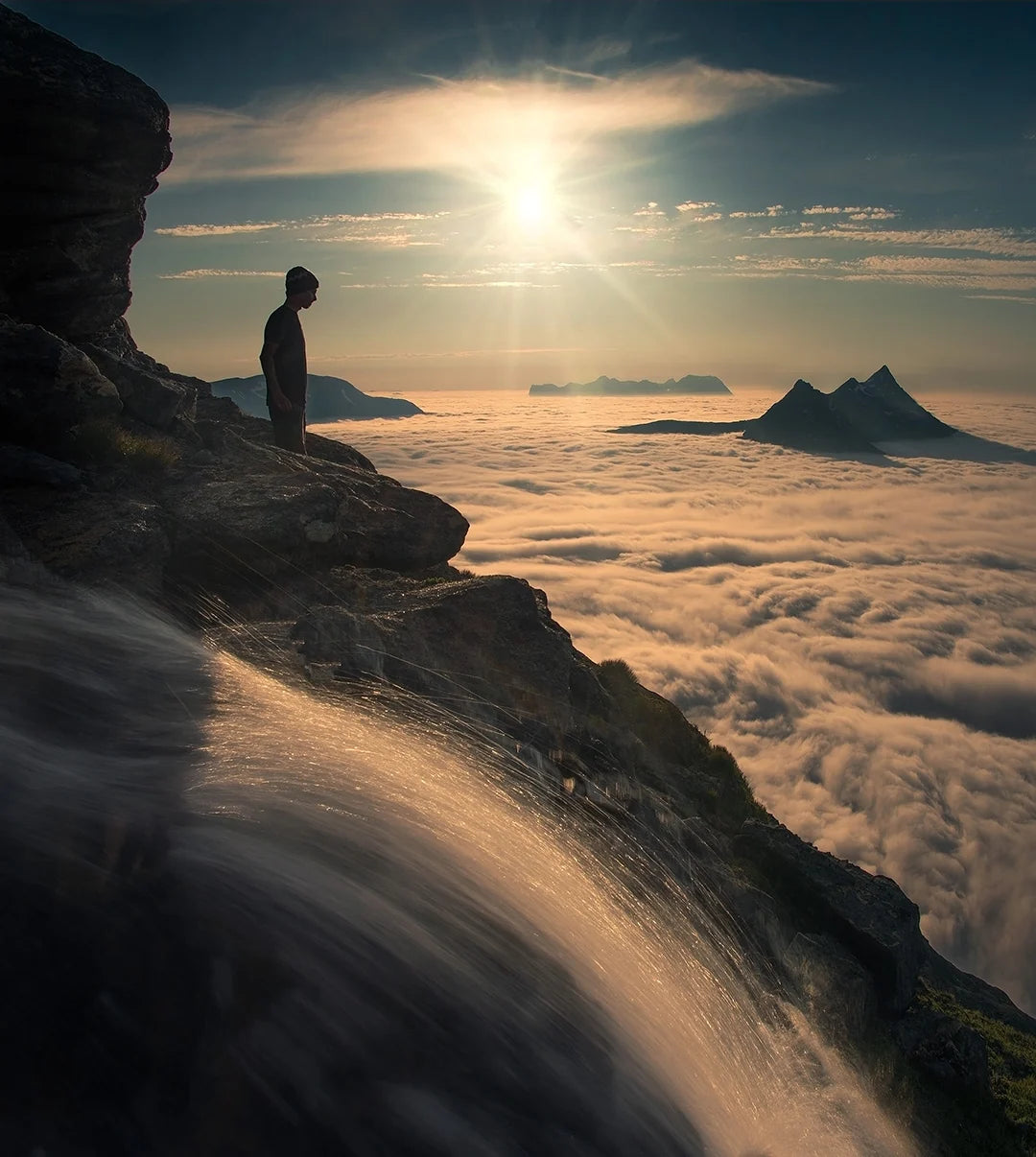 A person standing on a mountain ledge above a sea of clouds in golden light