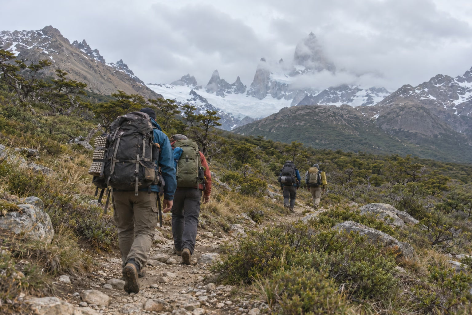 Four porters carrying large backpacks along a rocky trail beneath the mountains in Patagonia.