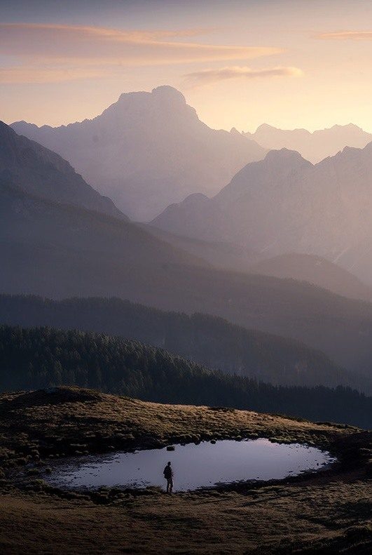 A small figure beside an alpine pool beneath layered mountain ridges in warm Dolomites light.