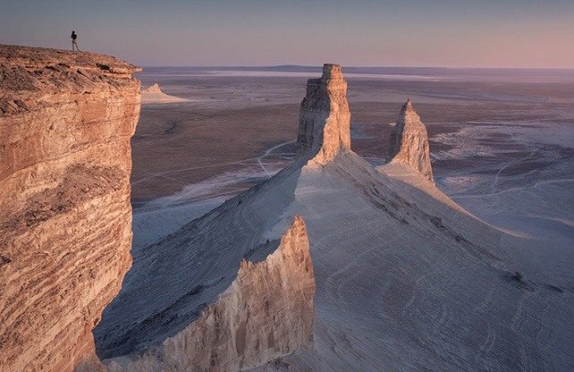 A person at the edge of a sunlit cliff overlooking pale desert pinnacles in Mangystau.