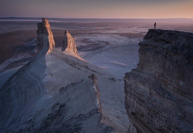 A figure on a dark cliff facing two pale pinnacles across the desert in Mangystau.