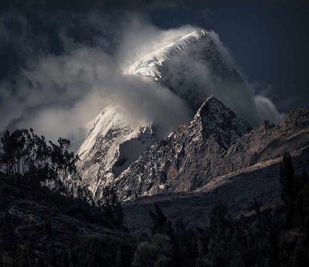 A snowy Peruvian peak emerging through cloud above dark trees and rocky slopes.