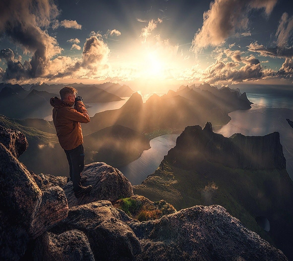 A photographer on a rocky summit as sunrays spread across Senja's mountains and fjords.