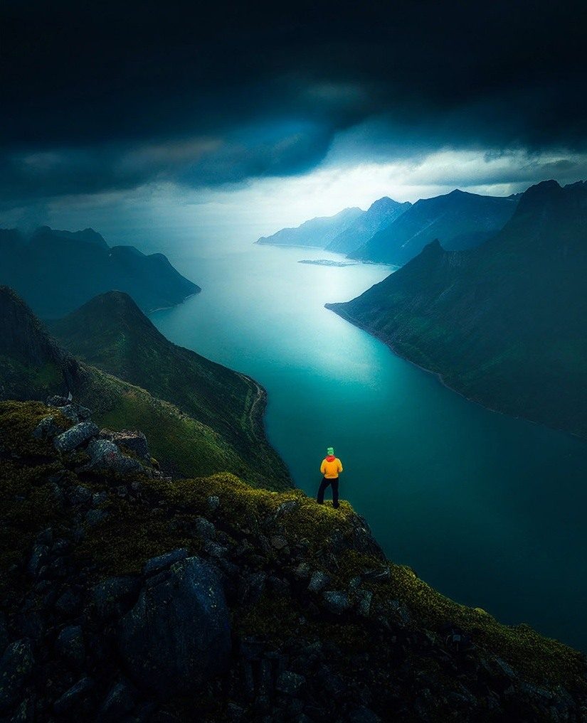 A hiker in a yellow jacket overlooking a turquoise fjord beneath dark clouds on Senja.